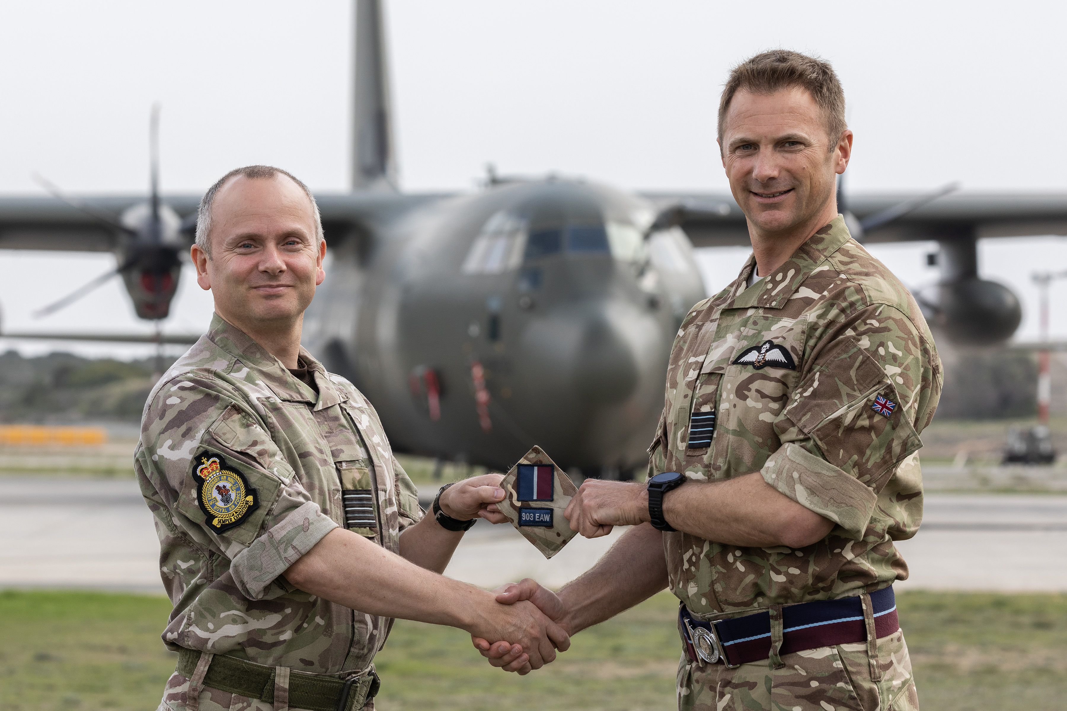Image shows RAF aviators shaking hands and holding Expeditionary Air Wing badge, in front of an Atlas aircraft.